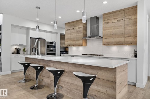 Contemporary kitchen featuring a large island with a white countertop and wood-finish paneling, stainless steel appliances, a white subway tile backsplash, and light wood-finish flooring - 1313 Hainstock Way Sw, Edmonton, AB - Indoor Photo Showing Kitchen With Upgraded Kitchen