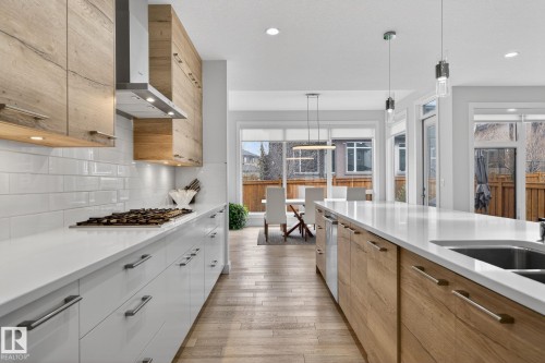 Gourmet kitchen featuring a gas cooktop, stainless steel range hood, contemporary wood-finish upper cabinetry, white lower cabinetry, and an expansive island with an undermount sink - 1313 Hainstock Way Sw, Edmonton, AB - Indoor Photo Showing Kitchen With Double Sink With Upgraded Kitchen