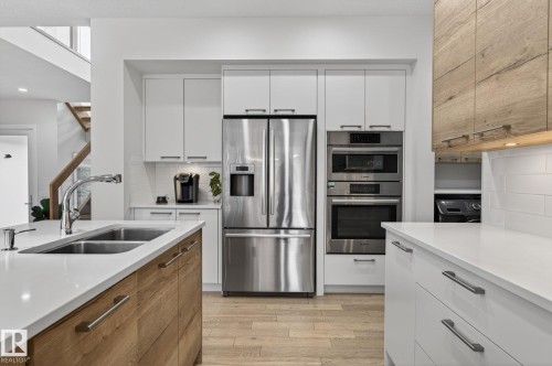 Modern kitchen featuring a double basin sink with a chrome faucet, stainless steel appliances, white cabinetry with silver hardware, light wood-finish flooring, and a subway tile backsplash - 1313 Hainstock Way Sw, Edmonton, AB - Indoor Photo Showing Kitchen With Double Sink With Upgraded Kitchen