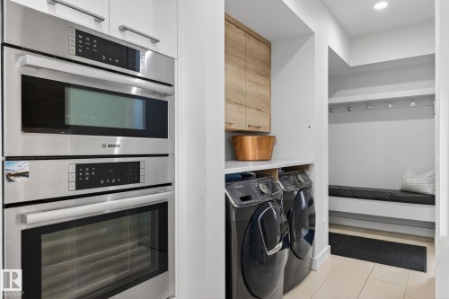 Built-in stainless steel oven and microwave combination unit paired with a dedicated laundry area featuring a washer and dryer, wood-finish cabinetry, and a mudroom-style bench with coat hooks - 1313 Hainstock Way Sw, Edmonton, AB - Indoor Photo Showing Laundry Room