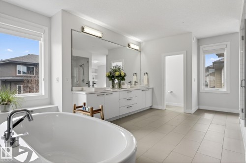 Freestanding oval soaking tub with chrome faucet, dual vanity with undermount sinks and quartz-style countertops, large wall-mounted mirror with overhead lighting, and tiled flooring - 1313 Hainstock Way Sw, Edmonton, AB - Indoor Photo Showing Bathroom