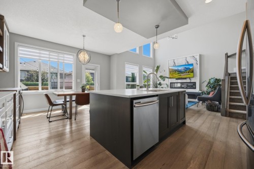 Open-concept living space featuring wood-finish flooring, a kitchen island with integrated stainless steel dishwasher, a wall-mounted fireplace with a marble-look surround, and a staircase with a contemporary wood and metal railing - 3870 Robins Crescent Nw, Edmonton, AB - Indoor Photo Showing Kitchen With Upgraded Kitchen