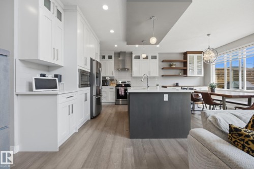 Open-concept kitchen featuring white shaker cabinetry, stainless steel appliances, a dark-toned island with a white countertop, wood-finish flooring, and a dining area with expansive windows - 3870 Robins Crescent Nw, Edmonton, AB - Indoor Photo Showing Kitchen With Upgraded Kitchen