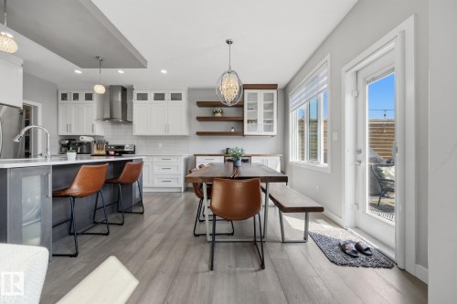 Open-concept kitchen and dining area featuring light wood-finish flooring, white cabinetry, a central island with integrated beverage cooler, a stainless steel range hood, and a patio door providing direct outdoor access - 3870 Robins Crescent Nw, Edmonton, AB - Indoor