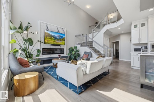Open-concept living space featuring high ceilings, wood-finish flooring, a modern fireplace with a marble-style surround, a contemporary staircase with glass railing, and a kitchen island with built-in storage - 3870 Robins Crescent Nw, Edmonton, AB - Indoor