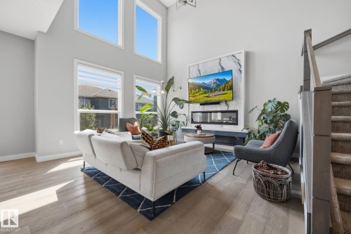 Spacious living area featuring high ceilings, tall windows, wood-finish flooring, a contemporary fireplace with a stone-look surround, and an adjacent staircase with carpeted treads - 3870 Robins Crescent Nw, Edmonton, AB - Indoor Photo Showing Living Room