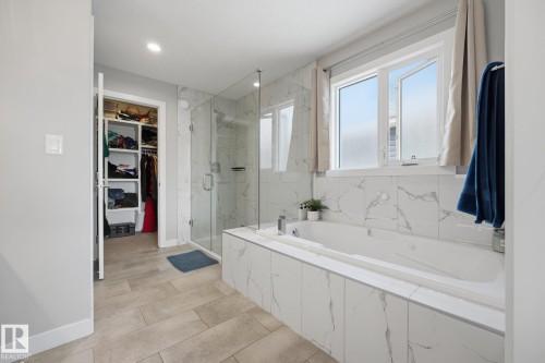Bathroom featuring a built-in soaking tub with marble-look tile surround, a glass-enclosed shower, dual-pane windows, and light wood-finish flooring - 3870 Robins Crescent Nw, Edmonton, AB - Indoor Photo Showing Bathroom
