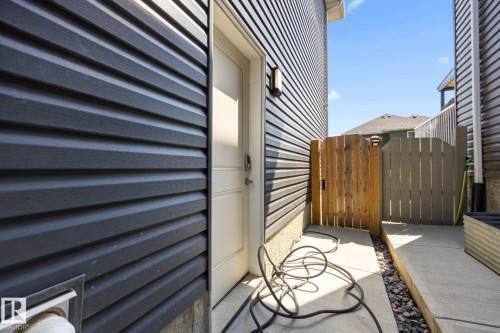 Exterior access area featuring dark horizontal siding, a light-colored entry door, a wrought iron wall sconce, and a wood privacy gate - 3870 Robins Crescent Nw, Edmonton, AB - Outdoor With Exterior