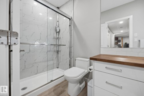 Bathroom featuring a glass-enclosed shower with large format marble-look tile, a white toilet, and wood-finish flooring - 3870 Robins Crescent Nw, Edmonton, AB - Indoor Photo Showing Bathroom