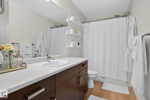 Bathroom featuring a dark wood-finish vanity with a white countertop and an integrated sink, a large wall-mounted mirror, a shower with a white curtain, and wood-finish flooring - #203 5521 7 Av Sw, Edmonton, AB - Indoor Photo Showing Bathroom