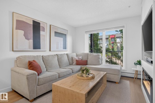 Bright living space featuring wood-finish flooring, a contemporary built-in entertainment unit, and a sliding glass door opening to an exterior balcony with black iron railings - #203 5521 7 Av Sw, Edmonton, AB - Indoor Photo Showing Living Room