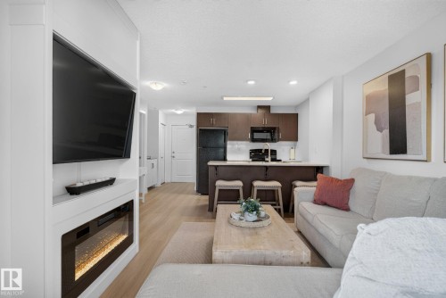Open-concept living space featuring wood-finish flooring, a built-in media console with an electric fireplace, dark wood cabinetry, and a kitchen island with seating - #203 5521 7 Av Sw, Edmonton, AB - Indoor Photo Showing Living Room With Fireplace