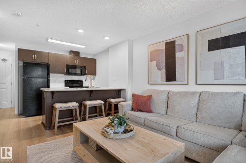 Open-concept living area featuring wood-finish flooring, a kitchen island with seating, dark wood cabinetry, and black appliances - #203 5521 7 Av Sw, Edmonton, AB - Indoor