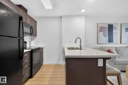 Kitchen featuring dark cabinetry, black appliances, light-toned countertops, and a subway tile backsplash - #203 5521 7 Av Sw, Edmonton, AB - Indoor Photo Showing Kitchen With Double Sink