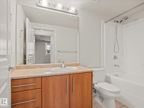 Bathroom featuring a wood-finish vanity with an integrated sink, a light-toned countertop, and a full-width mirror with overhead lighting - #707 9819 104 St Nw, Edmonton, AB - Indoor Photo Showing Bathroom