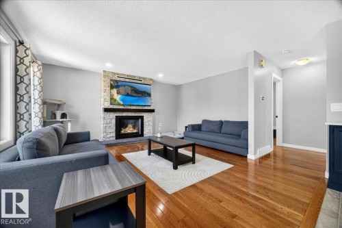 Living space featuring warm wood flooring, a stone-clad fireplace with a dark mantel, light gray wall finishes, a large window with patterned drapes, and a built-in dark blue cabinet - #47 21061 Wye Rd, Rural Strathcona County, AB - Indoor Photo Showing Living Room With Fireplace