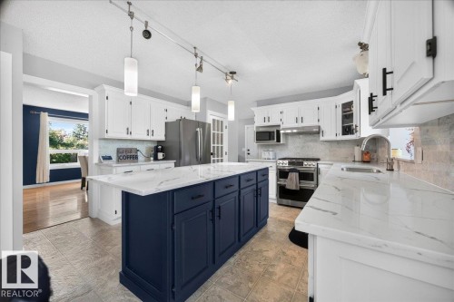 Spacious kitchen featuring a navy blue island, white perimeter cabinetry, stone-finish countertops, stainless steel appliances, and tile flooring - #47 21061 Wye Rd, Rural Strathcona County, AB - Indoor Photo Showing Kitchen With Upgraded Kitchen