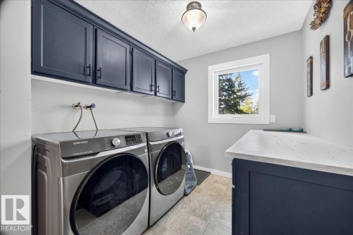 Laundry area featuring dark cabinetry with black hardware, a light countertop, a window with exterior tree scenery, and tile flooring - #47 21061 Wye Rd, Rural Strathcona County, AB - Indoor Photo Showing Laundry Room