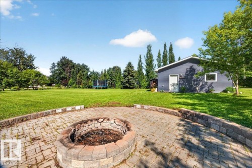 Paver patio with a stone-lined fire pit, surrounded by a large expanse of manicured lawn, mature trees, and an outbuilding featuring horizontal siding and a white entry door - #47 21061 Wye Rd, Rural Strathcona County, AB - Outdoor
