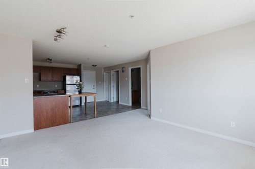 Open-concept living space featuring light-toned carpet flooring and a kitchen area with wood cabinetry, stainless steel refrigerator, and dark countertops - #7306 7327 South Terwillegar Dr Nw, Edmonton, AB - Indoor