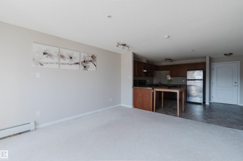 Open-concept living space featuring light-colored carpeting, a baseboard heater, and a kitchen with dark wood cabinetry, black countertops, and a stainless steel refrigerator - #7306 7327 South Terwillegar Dr Nw, Edmonton, AB - Indoor Photo Showing Kitchen