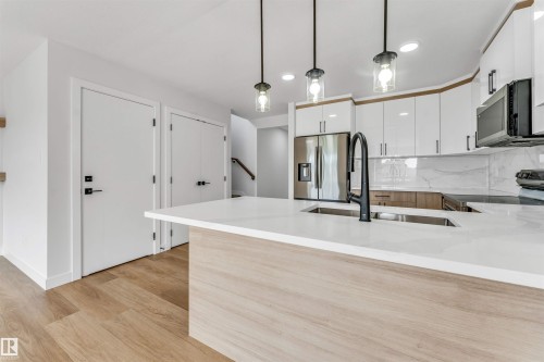 Kitchen featuring a large island with a double basin sink and matte black faucet, light wood-finish flooring, white flat-panel cabinetry, stainless steel appliances, and a marble-look backsplash - 1714 27 St. Nw, Edmonton, AB - Indoor Photo Showing Kitchen With Upgraded Kitchen