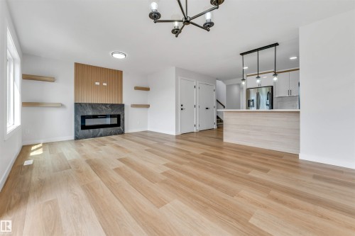 Spacious living area featuring light wood-finish flooring, a modern electric fireplace with a dark stone surround and wood-slat accent wall, and integrated shelving - 1714 27 St. Nw, Edmonton, AB - Indoor With Fireplace