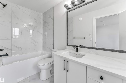 Contemporary bathroom featuring a white vanity with integrated sink, black fixtures, a framed mirror, and a built-in tub with white-veined tile surround - 1714 27 St. Nw, Edmonton, AB - Indoor Photo Showing Bathroom