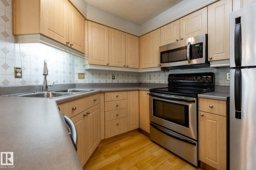 Kitchen featuring light wood-finish cabinetry with brushed nickel hardware, stainless steel appliances, a dual basin sink with a high-arc faucet, and wood-finish flooring - #103 12838 65 St Nw Nw, Edmonton, AB - Indoor Photo Showing Kitchen With Double Sink