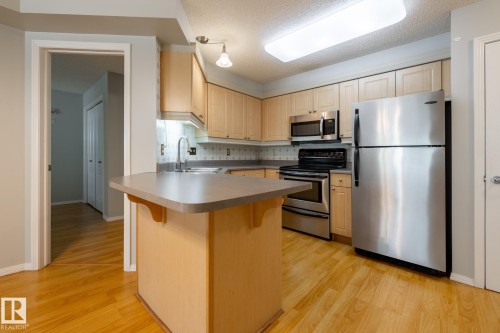 Kitchen featuring wood-finish flooring, light wood cabinetry, stainless steel appliances, a breakfast bar, and recessed lighting - #103 12838 65 St Nw Nw, Edmonton, AB - Indoor Photo Showing Kitchen
