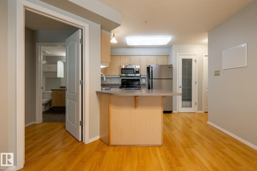 Kitchen featuring wood-finish flooring, light wood cabinetry, stainless steel appliances, a breakfast bar, and a tiled backsplash - #103 12838 65 St Nw Nw, Edmonton, AB - Indoor Photo Showing Kitchen