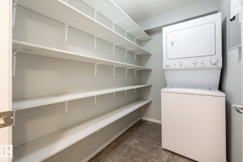 Dedicated utility space featuring a stacked washer and dryer unit, extensive wall-mounted shelving, neutral wall paint, and tiled flooring - #103 12838 65 St Nw Nw, Edmonton, AB - Indoor Photo Showing Laundry Room