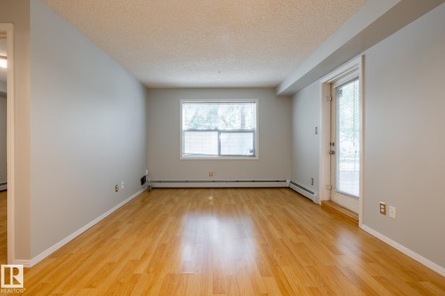 Spacious interior featuring wood-finish flooring, light grey painted walls, white trim, a large window, and a glass-paneled door - #103 12838 65 St Nw Nw, Edmonton, AB - Indoor Photo Showing Other Room