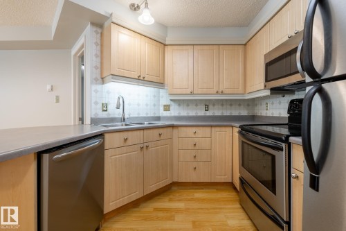 Kitchen featuring light wood cabinetry, stainless steel appliances, grey countertops, patterned tile backsplash, and wood-finish flooring - #103 12838 65 St Nw Nw, Edmonton, AB - Indoor Photo Showing Kitchen With Double Sink