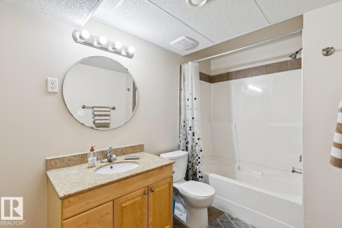 Bathroom featuring a wood-finish vanity with a granite-style countertop, a circular wall mirror with a four-bulb vanity light, and a white bathtub with a tiled surround - 16765 119 Street Nw, Edmonton, AB - Indoor Photo Showing Bathroom