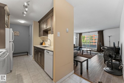 Open-concept living space with a kitchen featuring dark wood cabinetry and a white refrigerator, complemented by a living area with wood-finish flooring and a large window overlooking exterior buildings - #503 10175 114 St Nw, Edmonton, AB - Indoor Photo Showing Kitchen