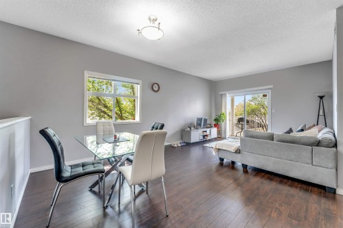 Open-concept living area featuring wood-finish flooring, light gray wall tones, a large window, and a sliding glass door providing outdoor access - #102 3040 Spence Wd Sw, Edmonton, AB - Indoor