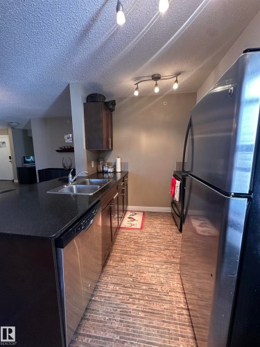 Kitchen featuring dark cabinetry, dark countertops, stainless steel appliances, and wood-finish flooring - #419 11808 22 Av Sw, Edmonton, AB - Indoor Photo Showing Kitchen With Double Sink
