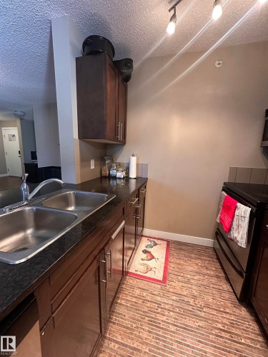 Kitchen featuring dark wood-finish cabinetry, a double basin stainless steel sink, black countertops, and a full-size black range - #419 11808 22 Av Sw, Edmonton, AB - Indoor Photo Showing Kitchen With Double Sink