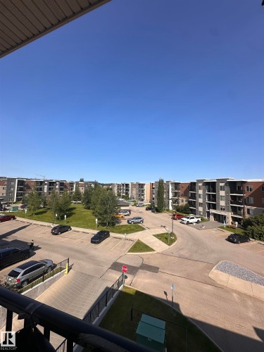 Panoramic outdoor scene featuring multiple residential buildings with varied facades, a central green space with mature trees, and a paved parking area with designated spaces - #419 11808 22 Av Sw, Edmonton, AB - Outdoor With View