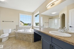 Spacious bathroom featuring a built-in soaker tub with tile surround, two large windows, a double vanity with a light-colored countertop and dark cabinetry, and a glass-enclosed shower - 