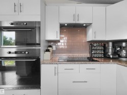 Contemporary kitchen featuring white cabinetry, stainless steel double wall ovens, a built-in cooktop, light-colored granite countertops, and a tiled subway backsplash - 