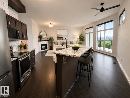 Open-concept living space featuring wood-finish flooring, a kitchen island with seating, a built-in fireplace, dark cabinetry, and expansive windows - #412 9503 101 Av Nw, Edmonton, AB - Indoor Photo Showing Kitchen With Upgraded Kitchen