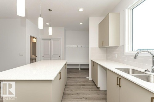 Kitchen featuring a large central island with light-toned countertops, contemporary pendant lighting, a dual-basin stainless steel sink with a gooseneck faucet, light-toned cabinetry with brass-finish hardware, and wood-finish flooring - 6269 175A Avenue Nw, Edmonton, AB - Indoor Photo Showing Kitchen With Double Sink