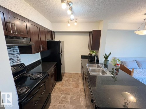 Kitchen featuring dark wood-finish cabinetry, black appliances, a mosaic tile backsplash, and dark countertops - #107 530 Watt Bv Sw, Edmonton, AB - Indoor Photo Showing Kitchen With Double Sink