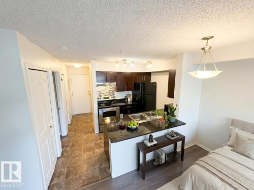 Open-concept kitchen featuring dark wood cabinetry, a mosaic tile backsplash, black countertops, and stainless steel appliances - #107 530 Watt Bv Sw, Edmonton, AB - Indoor