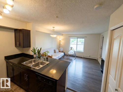 Kitchen island with an undermount double basin stainless steel sink, dark cabinetry, and dark countertops - #107 530 Watt Bv Sw, Edmonton, AB - Indoor Photo Showing Kitchen With Double Sink