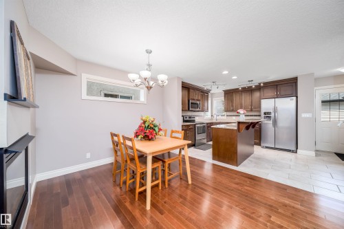 Expansive kitchen and dining area featuring wood-finish flooring, stainless steel appliances, dark wood cabinetry, a center island with countertop seating, and a built-in electric fireplace - 11342 79 Avenue, Edmonton, AB - Indoor