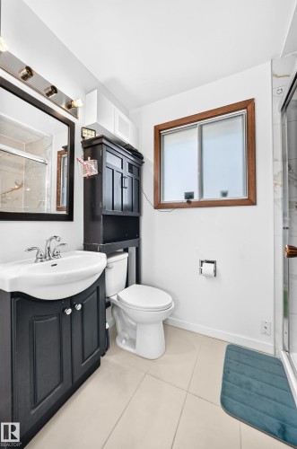 Bathroom featuring a dark wood-finish vanity with a white basin sink, a toilet with an adjacent dark wood-finish storage cabinet, a wood-framed window, and light-toned floor tiles - 1928 68 Street Nw, Edmonton, AB - Indoor Photo Showing Bathroom