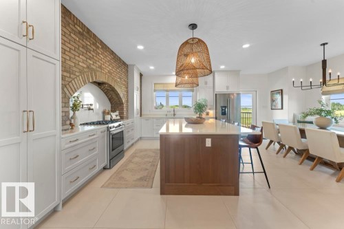 Spacious kitchen featuring an exposed brick archway, white cabinetry with champagne bronze hardware, a generous center island with a stone countertop, stainless steel appliances, and large windows - 2503 Lockhart Way, Cold Lake, AB - Indoor Photo Showing Kitchen With Upgraded Kitchen